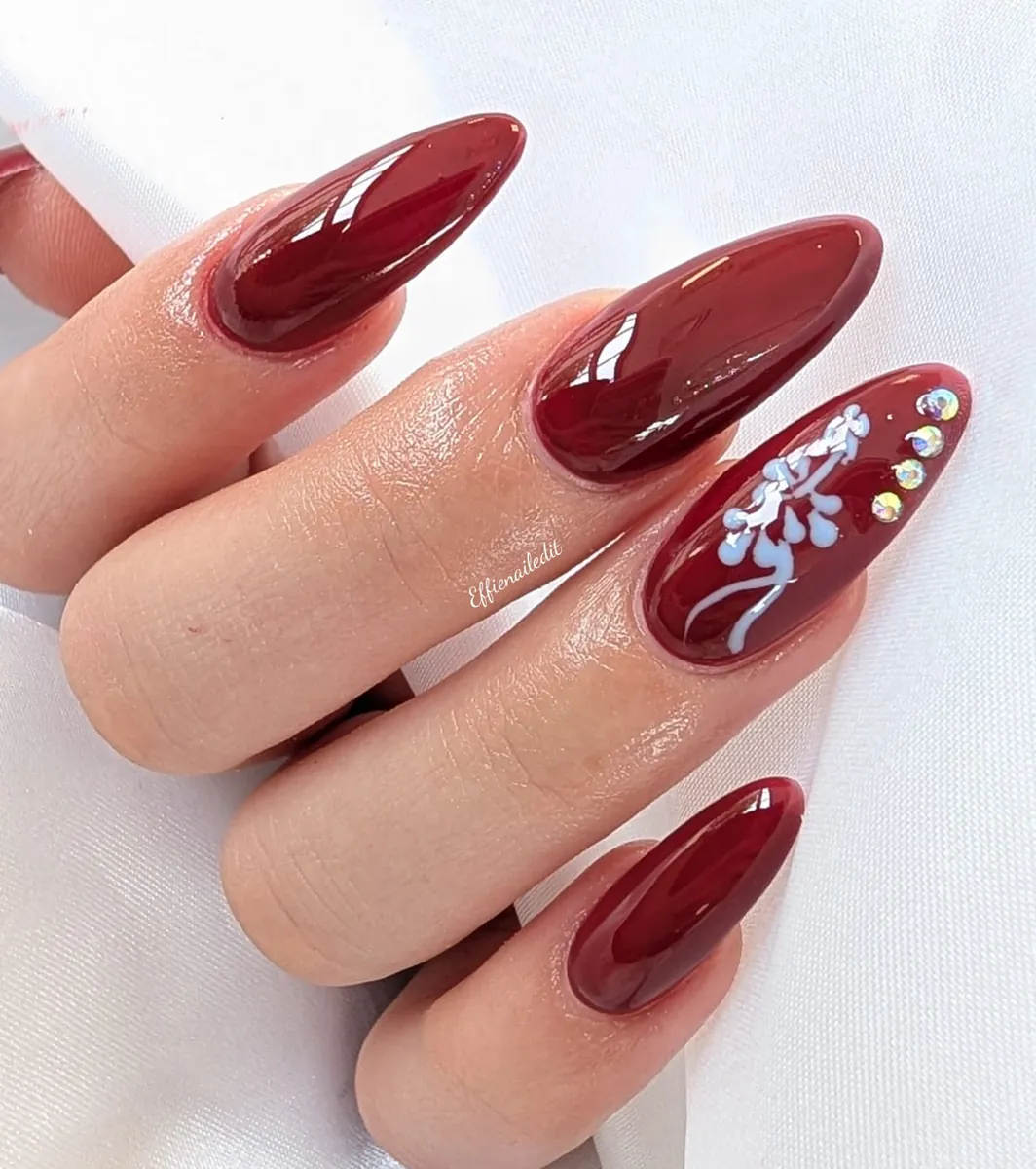 Glossy burgundy almond nails with a hand-painted floral accent and crystals