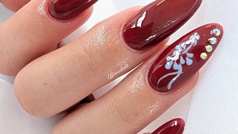 Glossy burgundy almond nails with a hand-painted floral accent and crystals