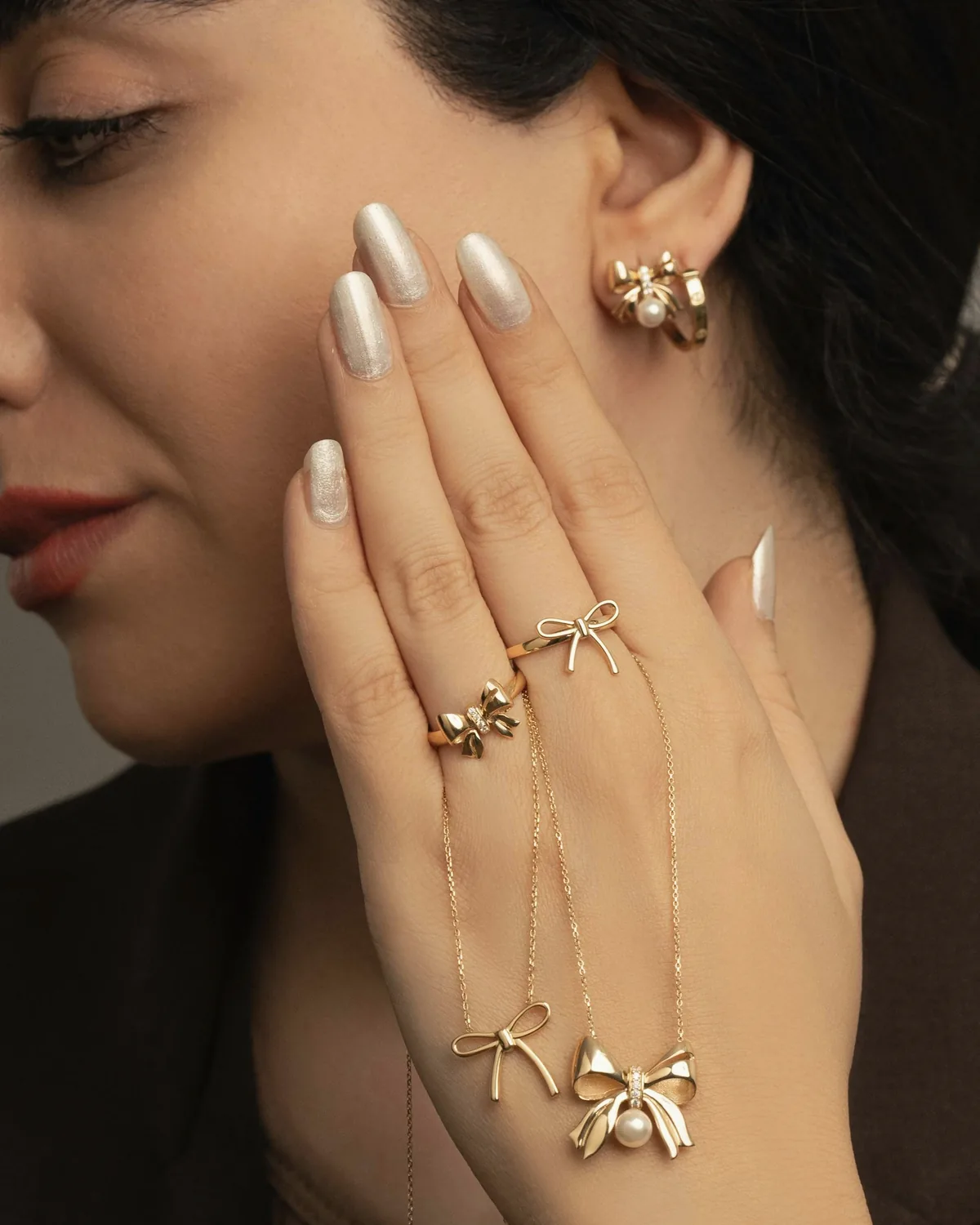 Pearl chrome almond nails with delicate gold rings, resting against her cheek